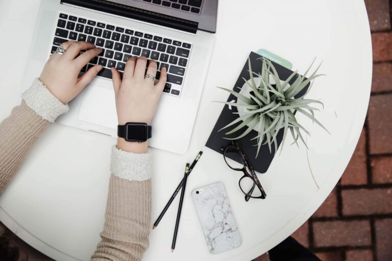 a person working on her laptop with spectacles, mobile, pencils, etc beside it