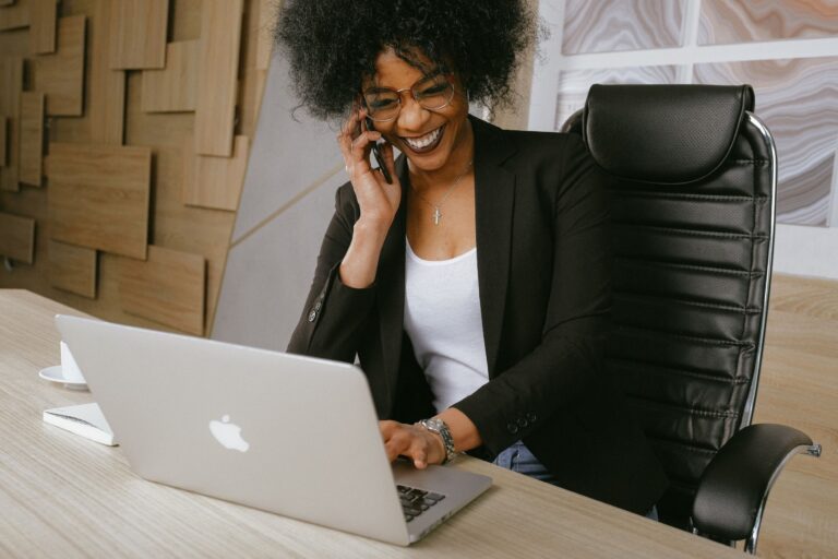 a woman smiling while talking on the phone in front of her laptop
