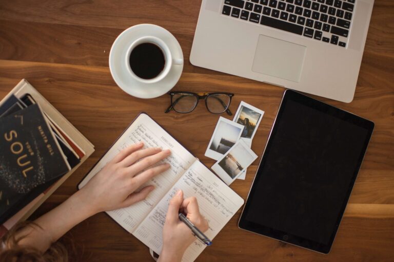 a person writing in a notebook on table surrounded by books, pictures, a tablet, spectacles, teacup and macbook