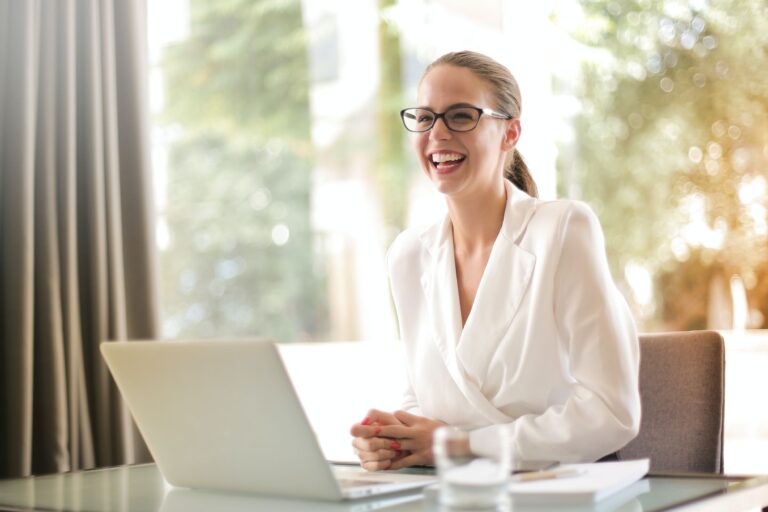 a woman sitting on a chair in a white suit with a laptop in front and smiling